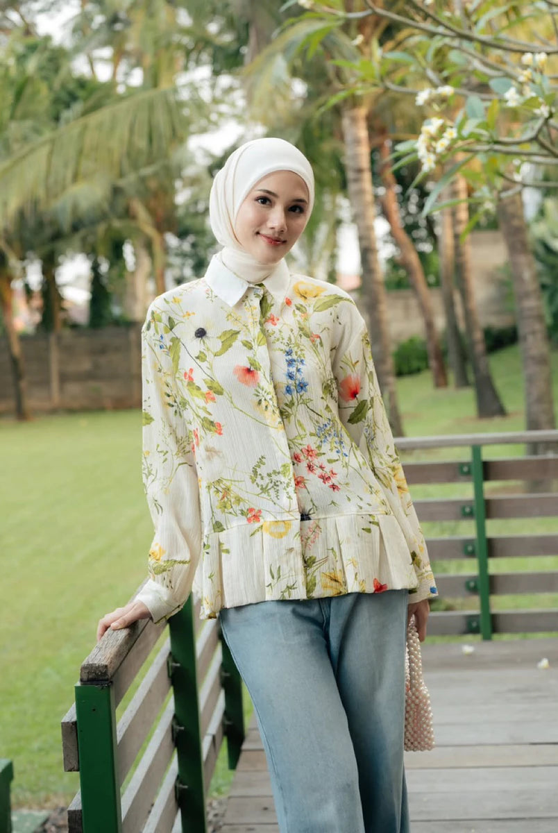 A woman wearing a cream floral long-sleeve blouse by Haidee Orlin with a white hijab and light blue jeans, standing on a wooden boardwalk in a park with green grass and palm trees in the background.