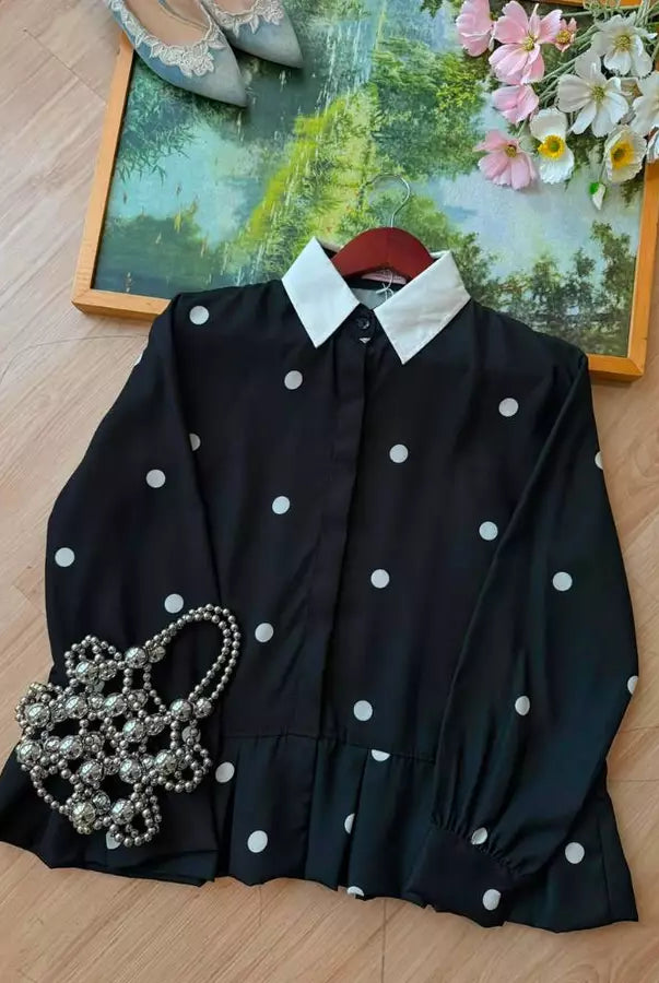 A black long-sleeve shirt by Haidee Orlin with white polka dots and a contrasting white pointed collar, laid flat on a light wood floor with decorative props including a pearl handbag, shoes, and a framed floral print.