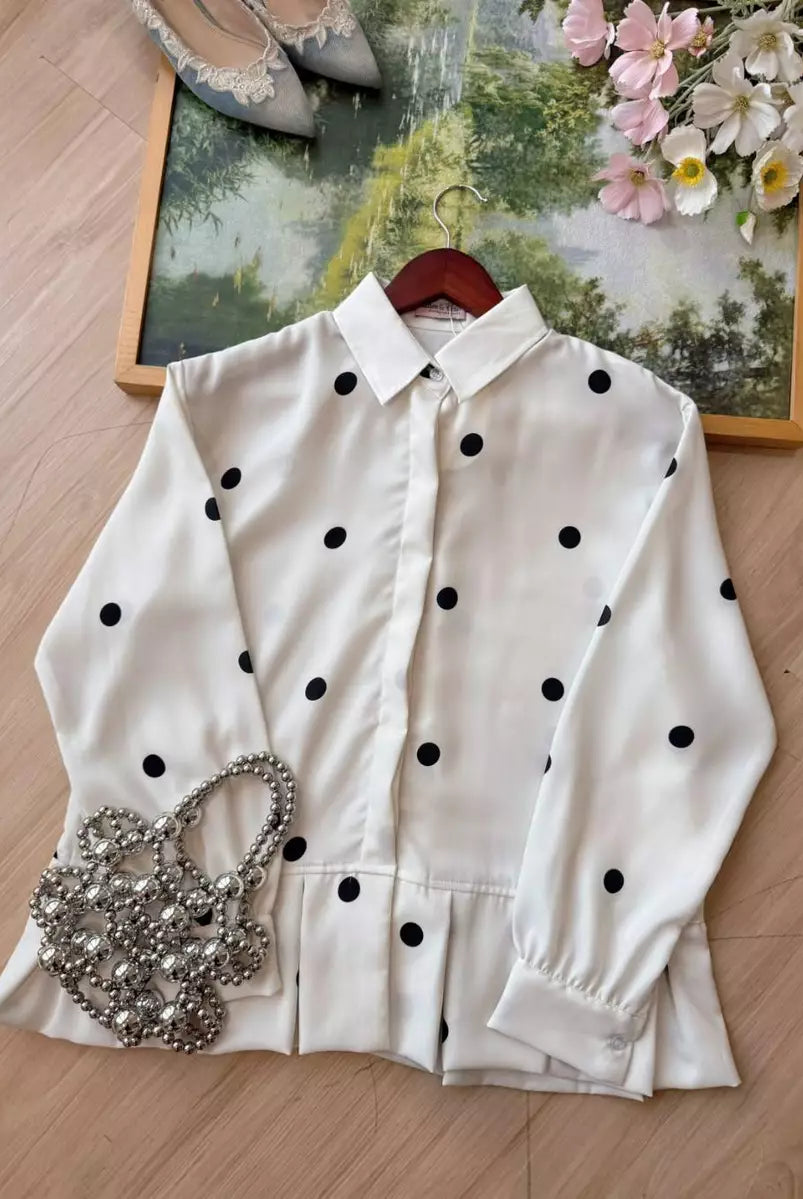 An off-white long-sleeve shirt by Haidee Orlin with black polka dots, styled on a wooden hanger on a light wood floor with a silver rhinestone purse, a pair of pointed shoes, and a floral picture frame in the background.