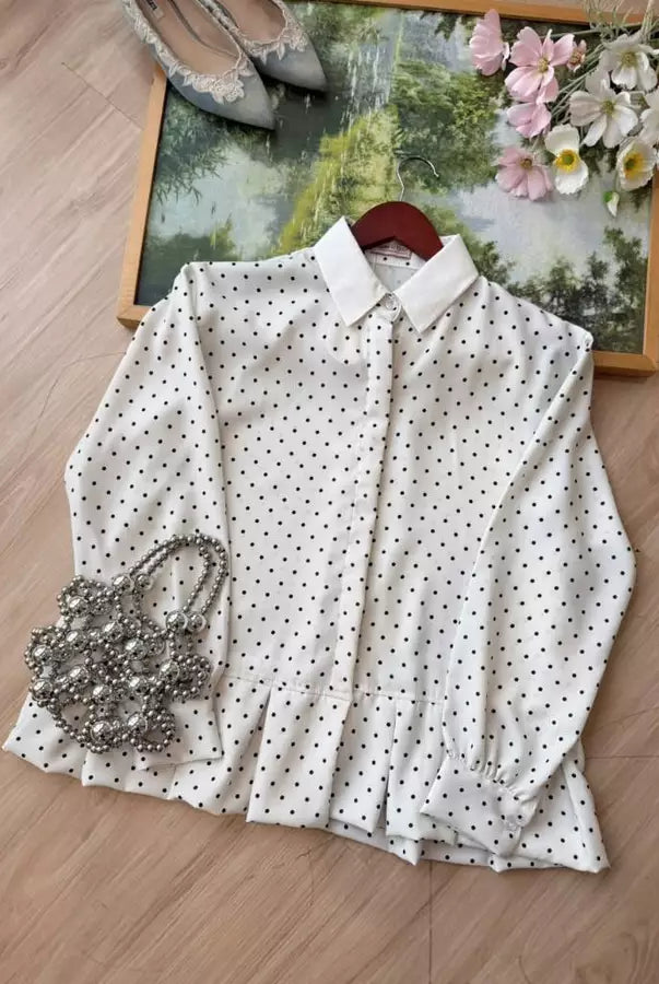 A white long-sleeve shirt by Haidee Orlin with a black polka dot pattern and a pointed collar, laid flat on a wooden floor with decorative accessories including a beaded handbag, shoes, and a framed picture.