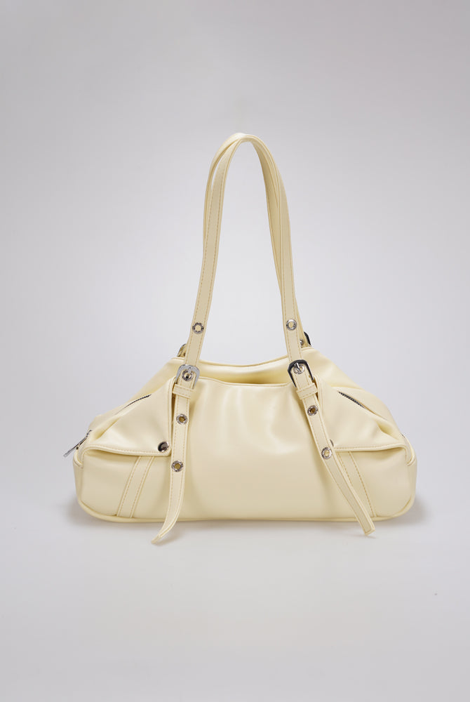 A cream-colored leather shoulder bag by Cekhas with a curved top handle, decorative buckle straps, and silver-tone hardware, shown against a neutral light gray background.