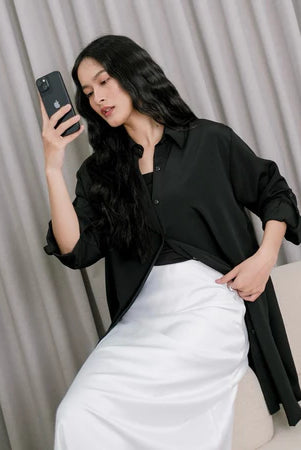 A woman wearing a black tunic over a black top and white pants, standing against a gray backdrop.