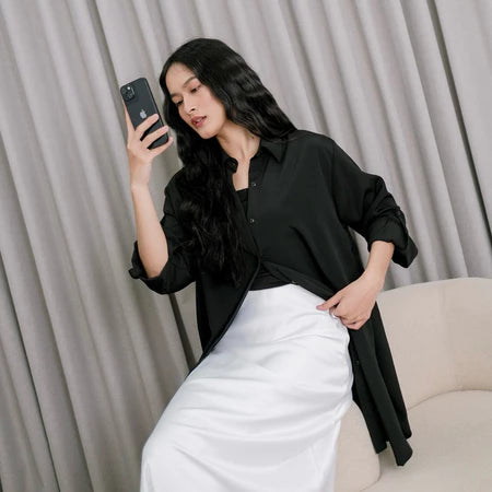 A woman wearing a black tunic over a black top and white pants, standing against a gray backdrop.