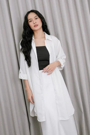 A woman wearing a white tunic over a black top and white pants, standing against a gray backdrop.