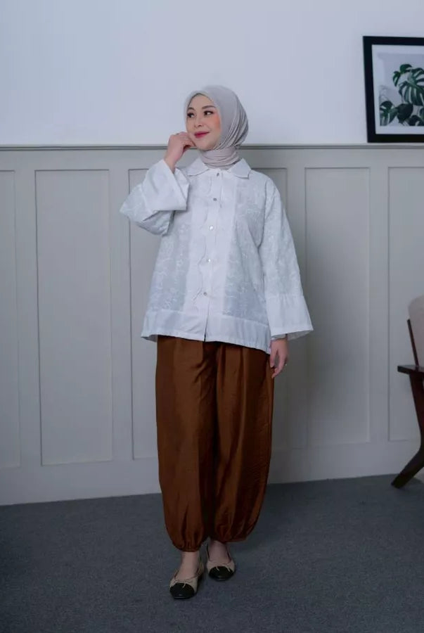 Woman wearing a white shirt by Abame and brown pants standing in a room with a white wall and a chair.