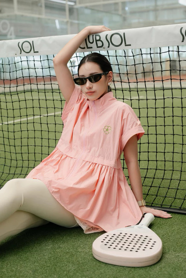 A woman wearing a light pink short-sleeve athletic vest by Haidee Orlin and beige leggings sits on a tennis court behind a net, holding a paddle. The dress has a small embroidered logo on the chest.