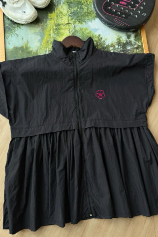A black sleeveless zip-front long vest by Haidee Orlin with a gathered waist and a small pink paw-print logo on the chest, laid flat on a wood floor with shoes and a bag partially visible around it.