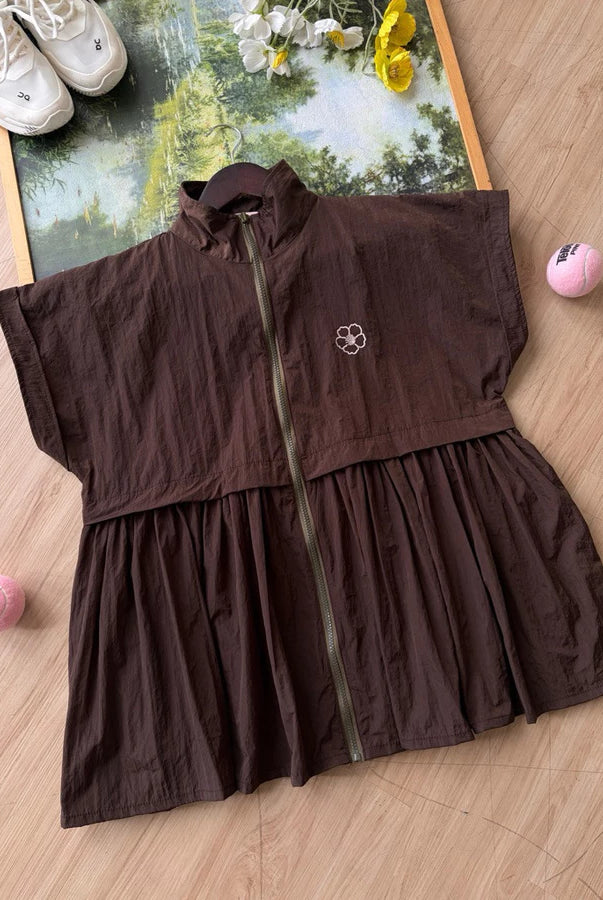 A dark brown sleeveless zip-front long vest by Haidee Orlin with a gathered waist seam, laid flat on a light wood floor. The garment has a small white embroidered logo on the chest and is styled with sneakers, flowers, and pink balls around it.