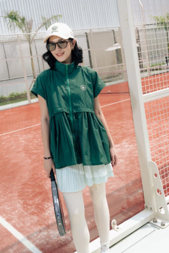A woman stands on a red clay tennis court wearing a dark green short-sleeve zip-front vest by Haidee Orlin over a light green pleated mini skirt, with white leggings, a white cap, and sunglasses, holding a tennis racket.