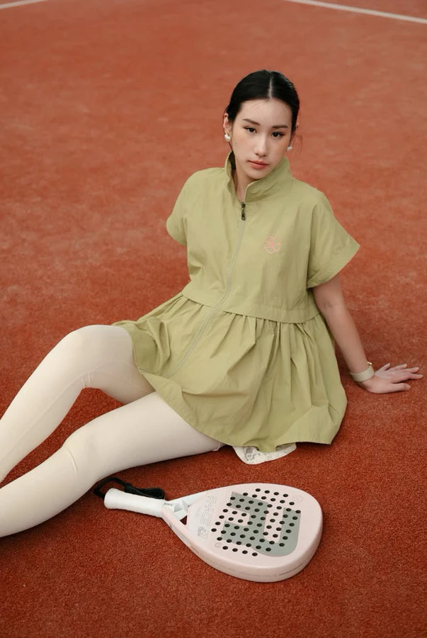A woman sits on a clay tennis court wearing a light olive green short-sleeve athletic vest by Haidee Orlin and white leggings, with a paddle racket in front of her.