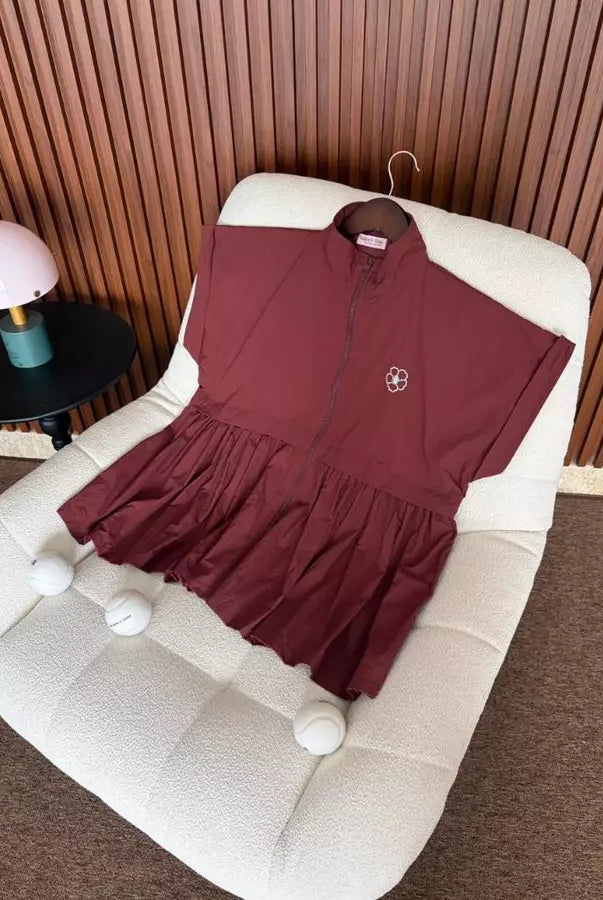 A burgundy short-sleeve zip-front longline vest by Haidee Orlin with a gathered ruffled hem, displayed on a cream boucle chair in an indoor setting with a slatted wood wall and a small table lamp.