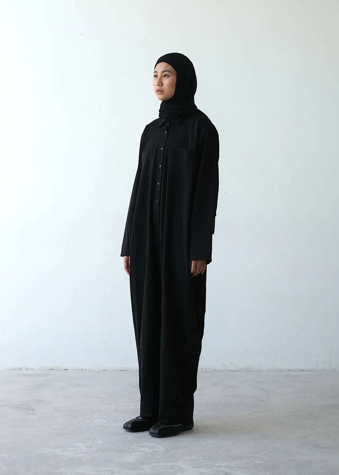 A person wearing a black hijab and a matching black jumpsuit by Rashawl with a button bustline, standing against a plain background.