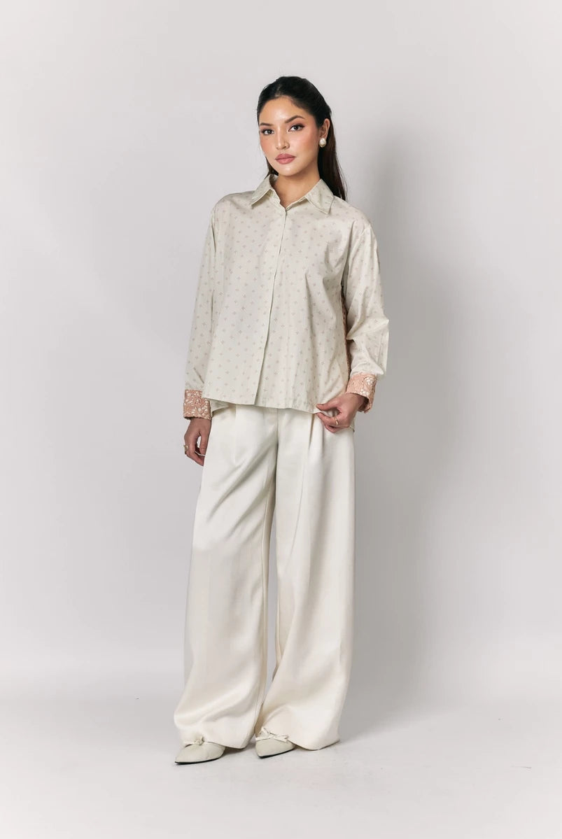 Woman wearing a beige shirt by Studio Tui and white pants against a plain background
