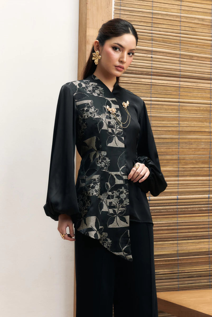 Woman wearing a black floral longsleeves blouse by Studio Tui against a wooden panel background