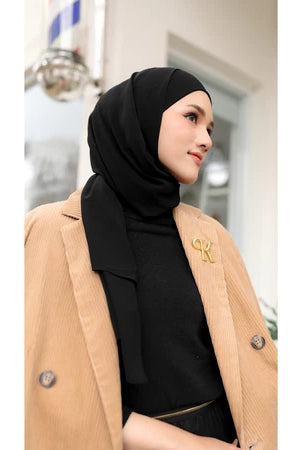 Woman wearing a black pashmina hijab by Puru Kambera and beige coat with a brand logo, standing outdoors.