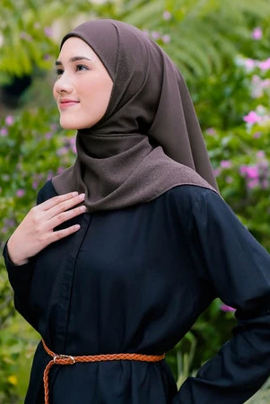 Woman wearing a brown pashmina hijab by Puru Kambera and black outfit with a belt, standing in a garden with greenery and flowers.