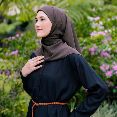 Woman wearing a brown pashmina hijab by Puru Kambera and black outfit with a belt, standing in a garden with greenery and flowers.
