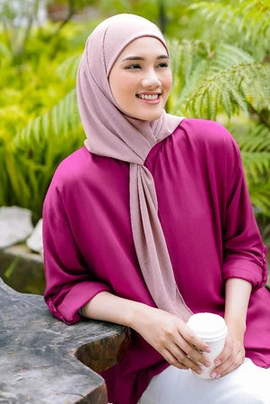 Woman in a pink outfit with a pashmina hijab by Puru Kambera sitting outdoors holding a white cup.