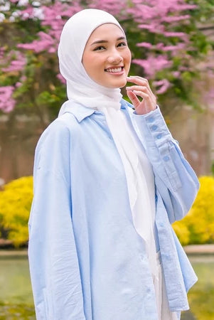 Woman wearing a light blue jacket and white pashmina hijab by Puru Kambera standing in front of pink flowers and greenery.