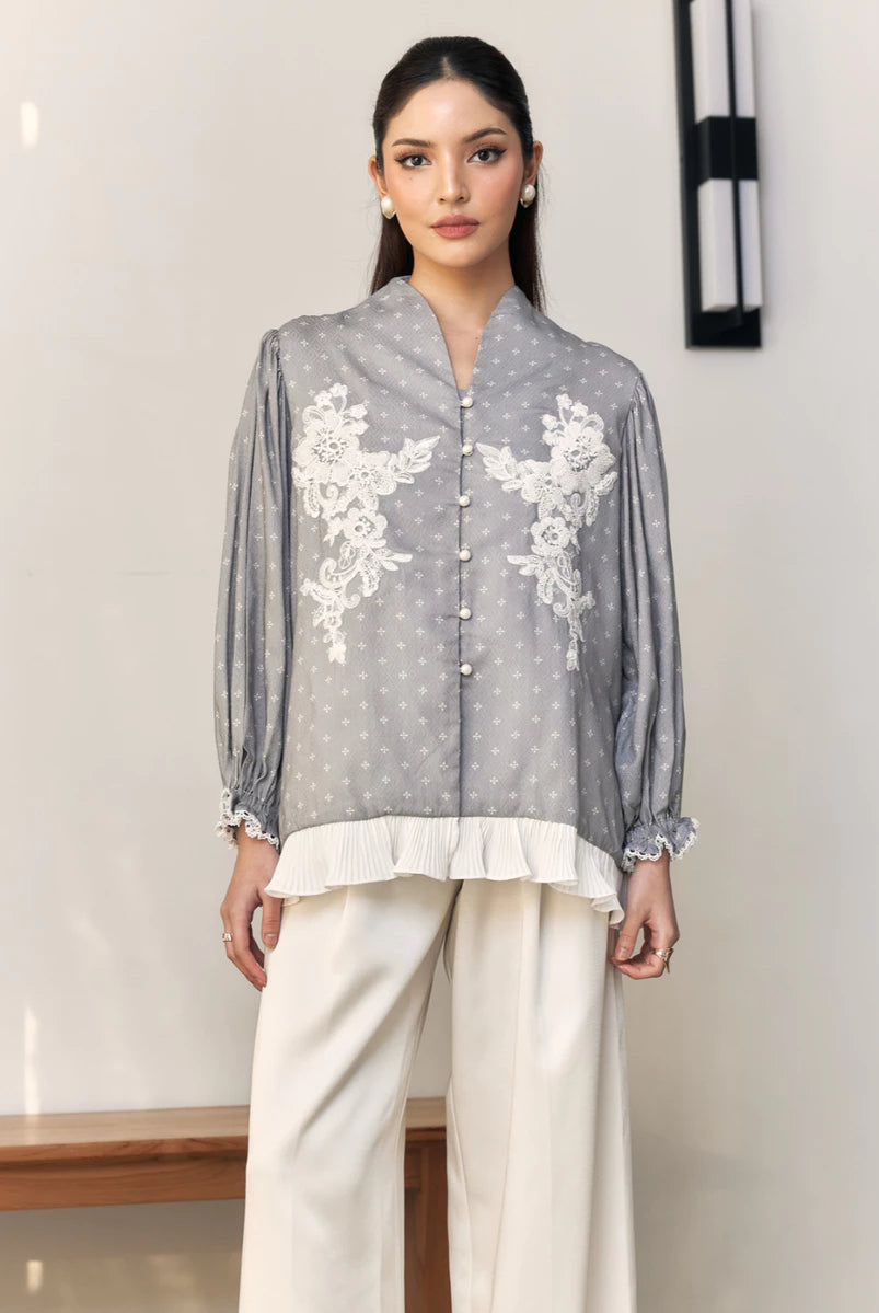 Woman wearing a gray blouse with white floral embroidery by Studio Tui and white pants indoors.