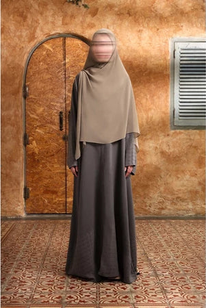 Person wearing a beige hijab and gray Mecca Dhurma Abaya by Nehhat Modest standing in front of a wooden door.