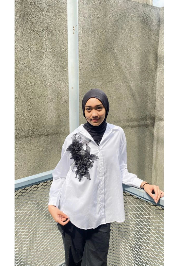 Person wearing a white shirt with black floral design from Maima and dark pants standing against a textured wall.