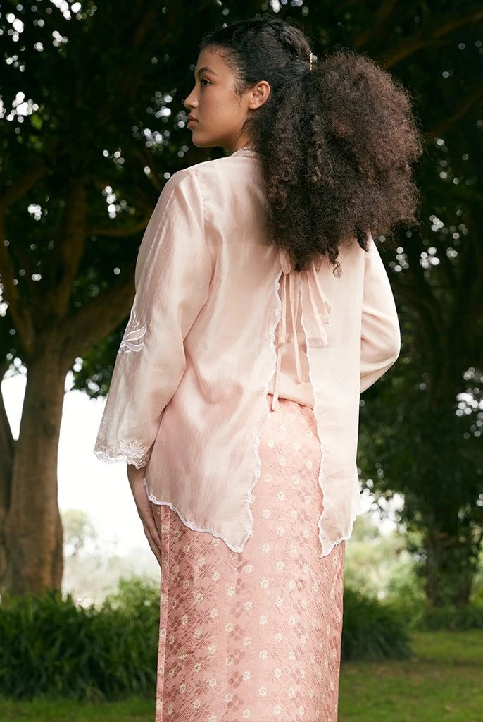 Woman in a peach colored outer by Kartinis Label standing in a park with trees in the background