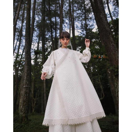A woman standing in a forest clearing wearing a white long-sleeved outer by Maima with a floral pattern and a V-neckline.