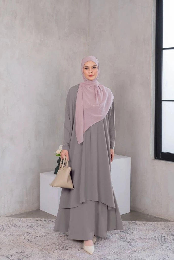 A woman wearing a dark brown tunic dress with a hijab, standing in a room with minimalist decor.