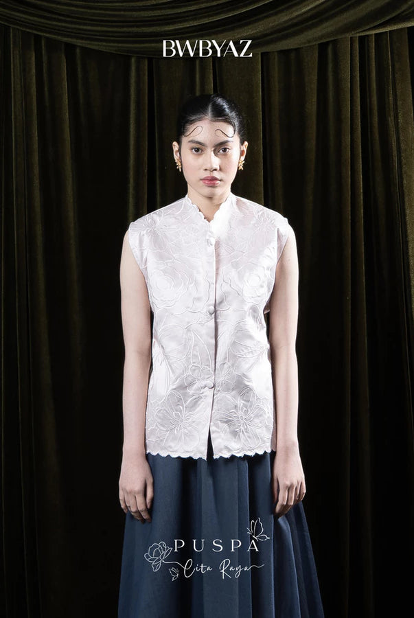Woman wearing a light pink sleeveless embroidered vest and dark skirt against a dark curtain background with 'BWBYAZ' branding.
