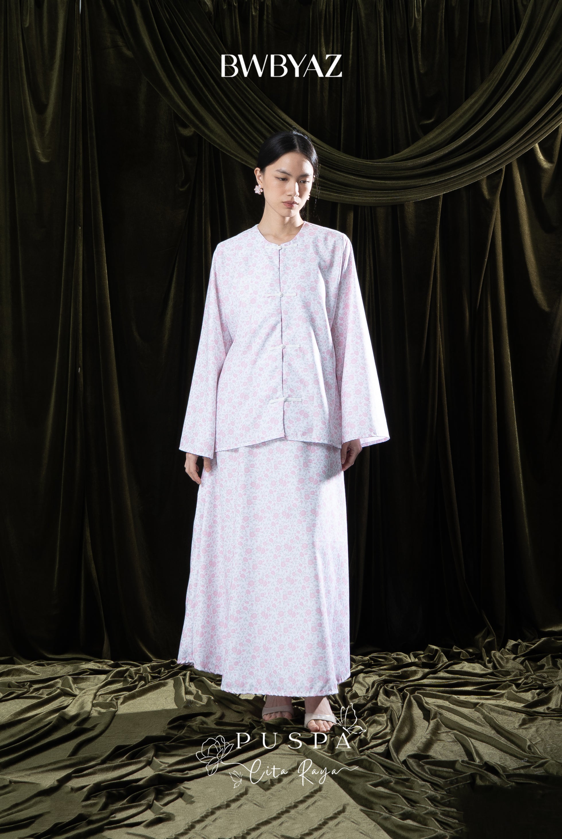 Woman wearing a light pink traditional outfit with 'BWBYAZ' branding against a dark curtain background.