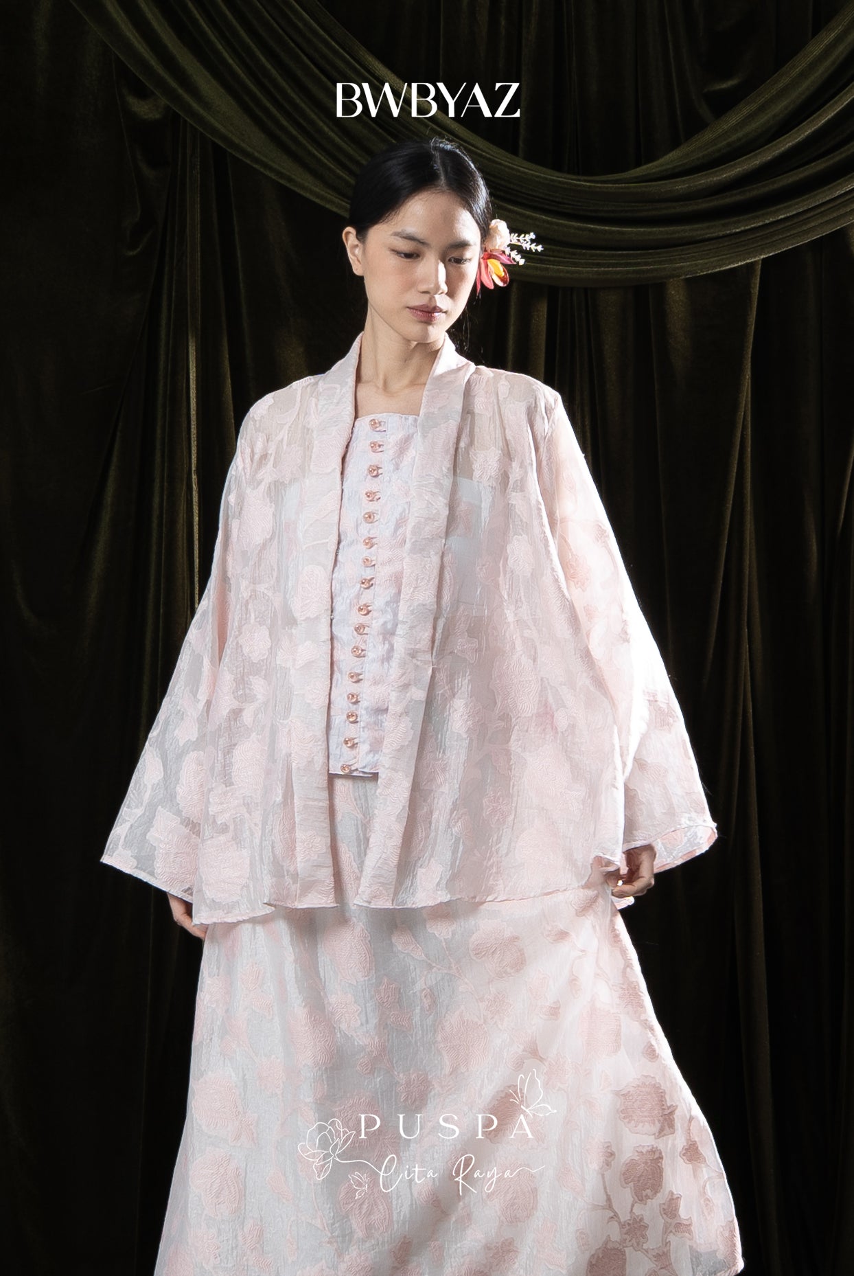 Woman wearing a light pink kebaya with lace details on a dark curtain background, featuring the brand 'BWBYAZ'.