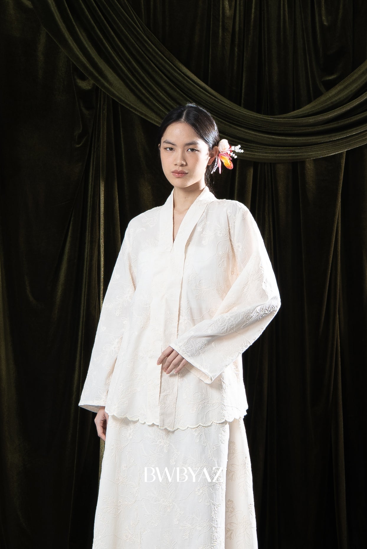 Woman in a cream traditional dress standing against a dark green curtain with 'BWBYAZ' branding.
Seorang wanita mengenakan kebaya berwarna krem berdiri di depan tirai hijau gelap dengan merek ‘BWBYAZ.