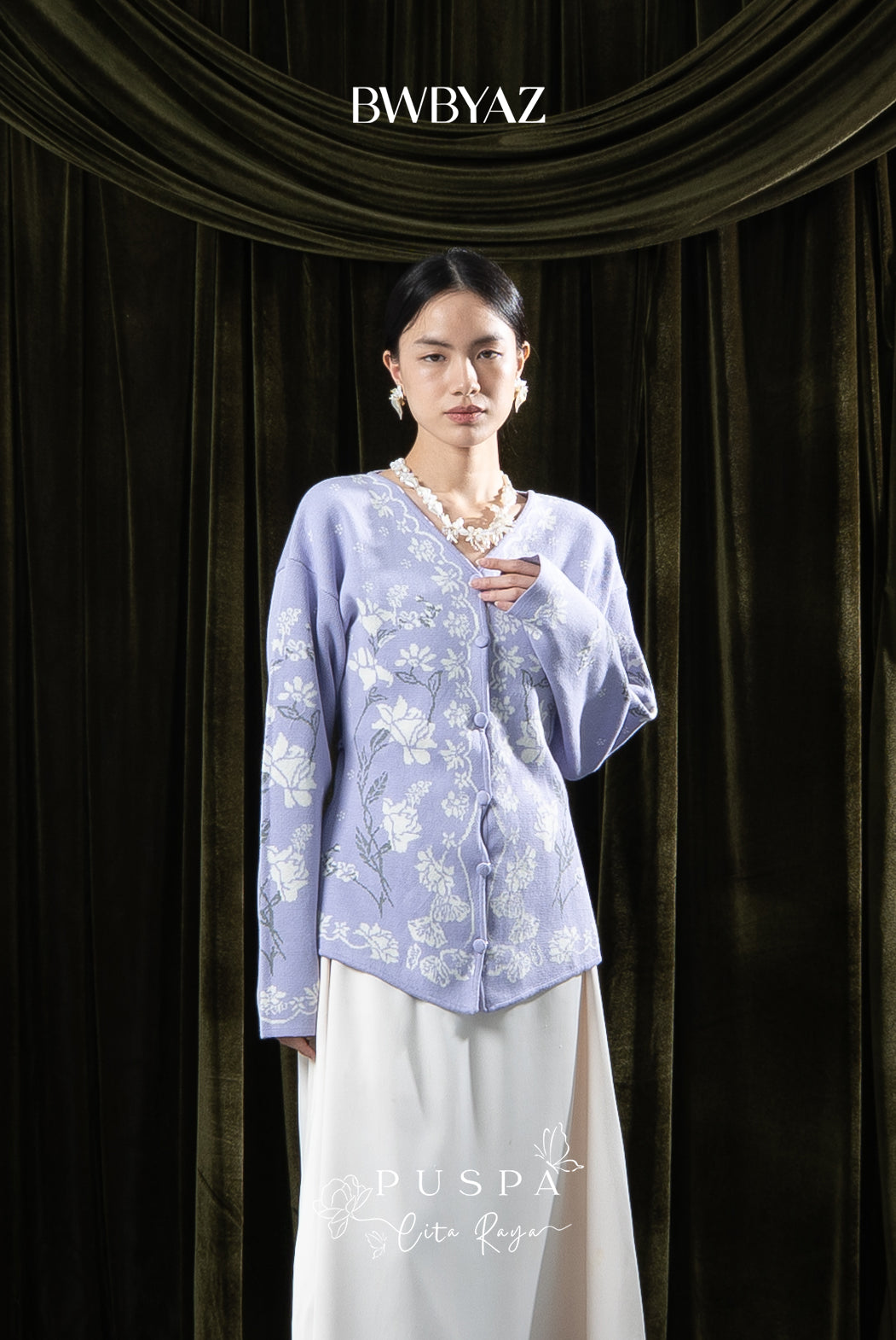 Woman wearing a lavender-colored floral cardigan and white skirt against a dark curtain backdrop with 'BWBYAZ' branding.
