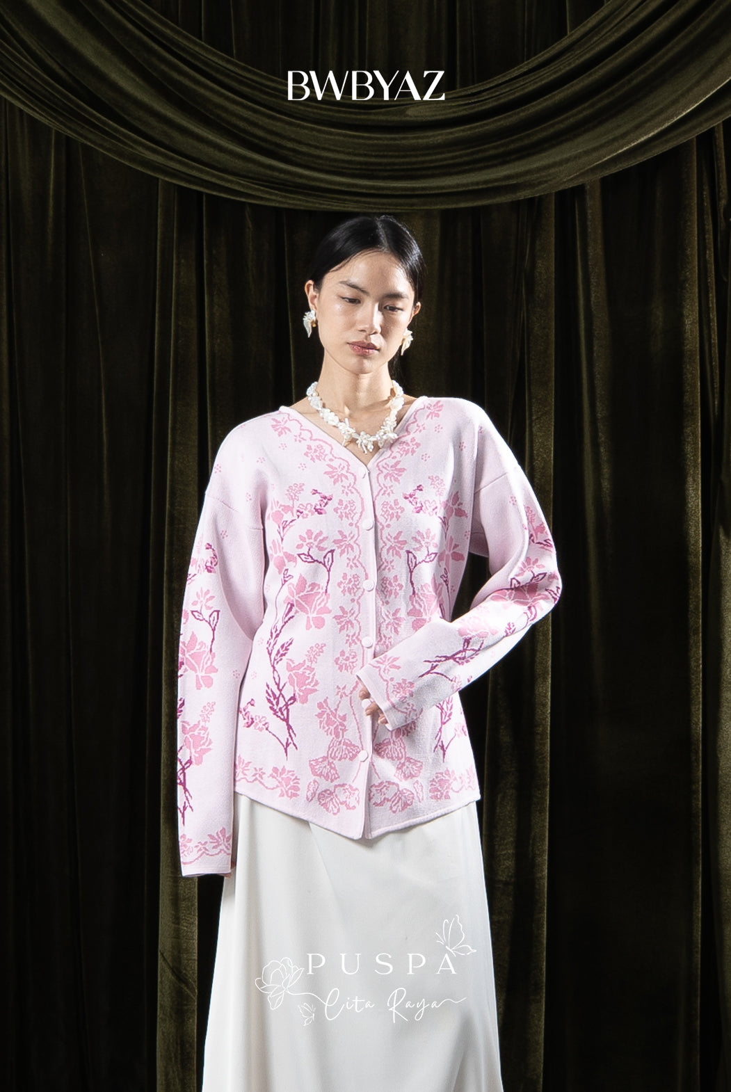 Woman wearing a pink floral cardigan and white skirt against a dark curtain backdrop with 'BWBYAZ' branding.