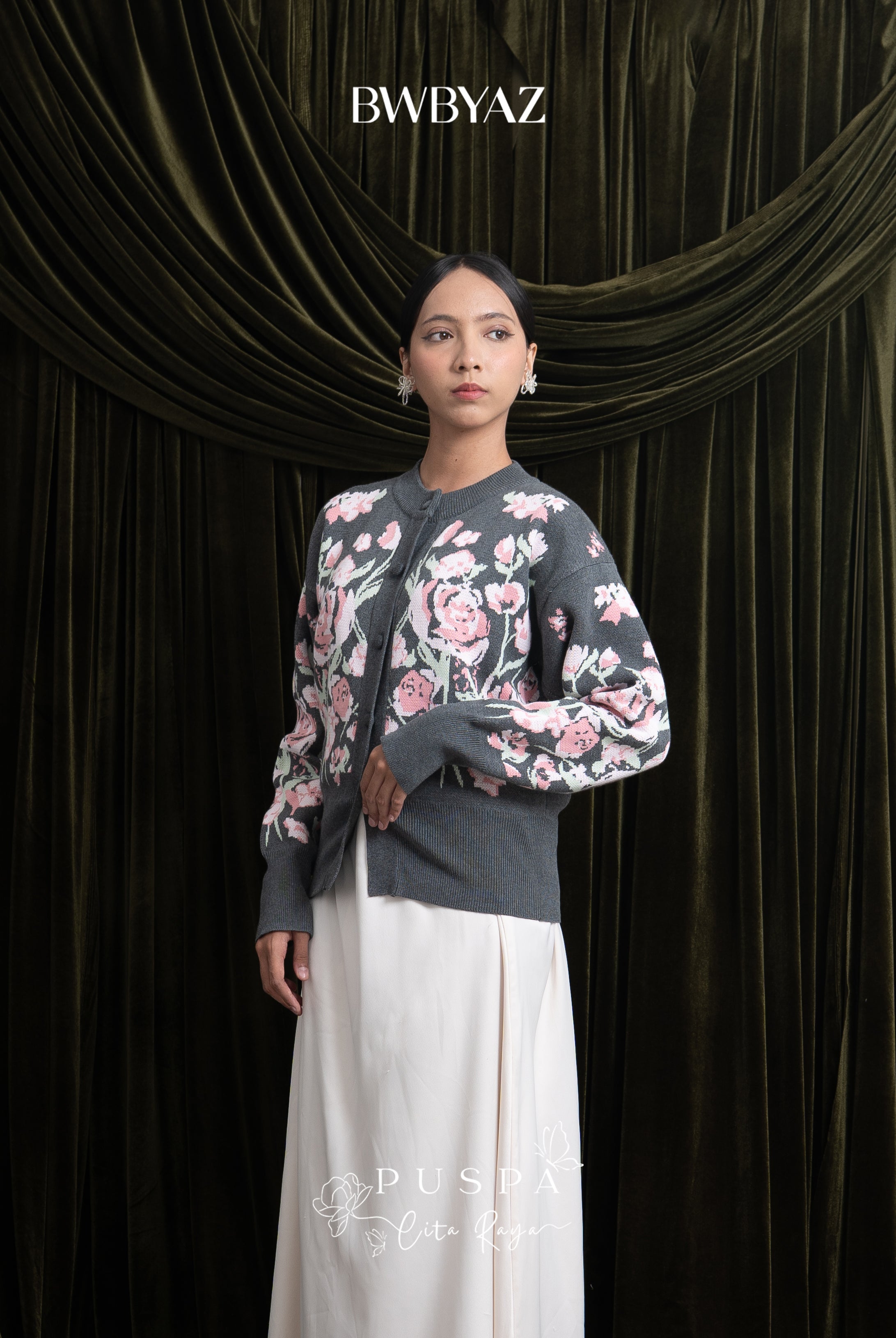 Woman wearing a grey floral cardigan and white skirt against a dark curtain backdrop with 'BWBYAZ' branding.