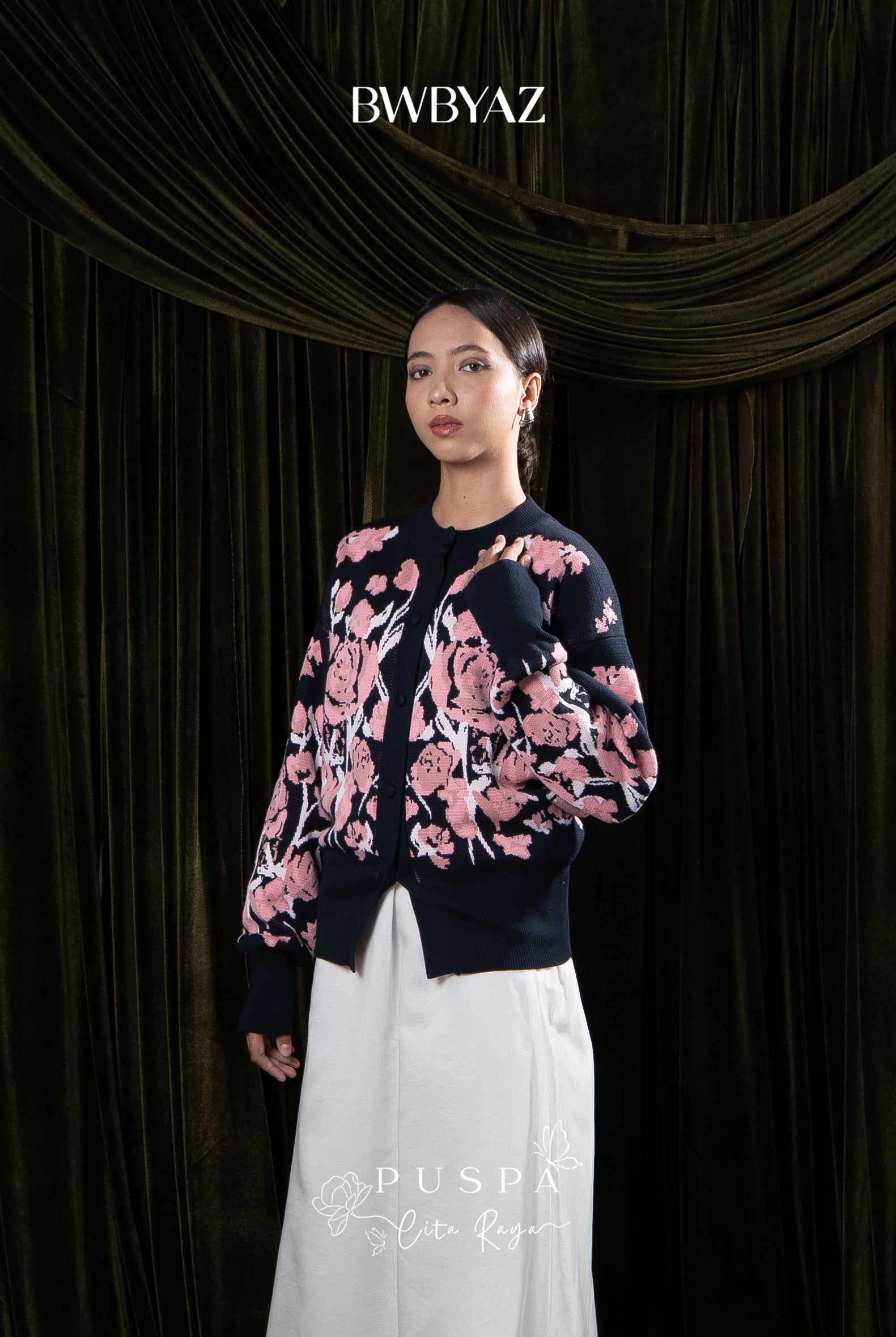 Woman wearing a navy floral cardigan and white skirt against a dark curtain background with 'BWBYAZ' branding.