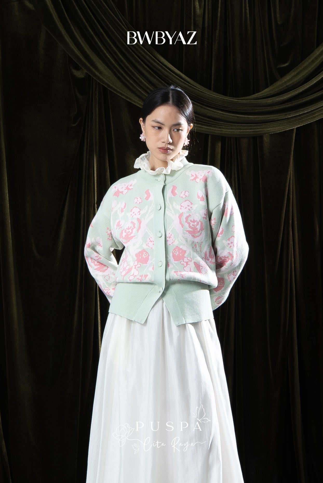 Woman wearing a light green floral cardigan with a white skirt against a dark curtain background, featuring the brand 'BWBYAZ'.