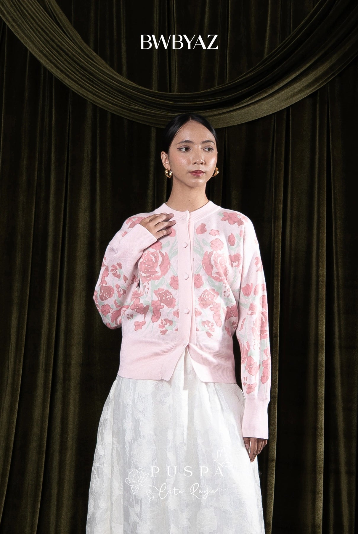 Woman wearing a pink floral cardigan and white skirt against a dark curtain background with 'BWBYAZ' branding.