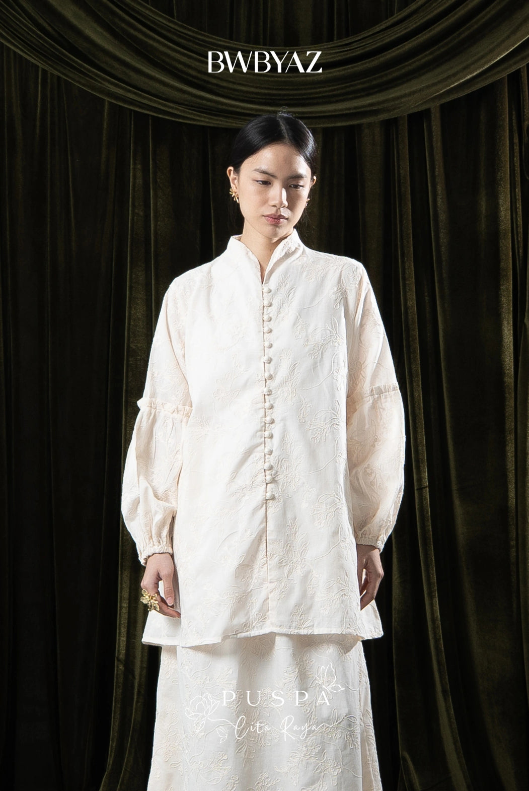 Woman wearing a cream tunic with lace details against a dark curtain backdrop with 'BWBYAZ' branding.
Seorang wanita mengenakan tunik krem dengan detail renda di depan latar belakang tirai gelap dengan merek ‘BWBYAZ’.