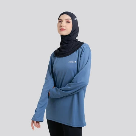Woman wearing a blue long-sleeve top by Noore and black hijab on a gray background