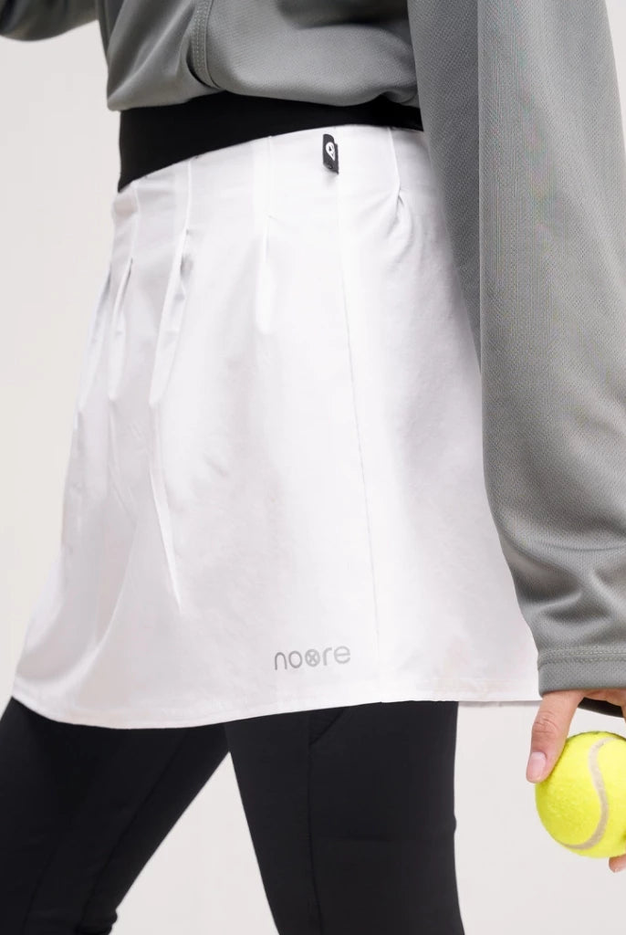 A person wearing a white tennis skirt by Noore with a logo on the lower left side, paired with black leggings and a gray sweatshirt. A tennis ball is held in the person's hand.