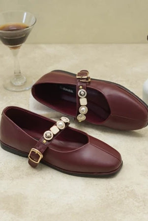 Pair of maroon flat shoes with decorative buckles by Dianable on a beige surface.
