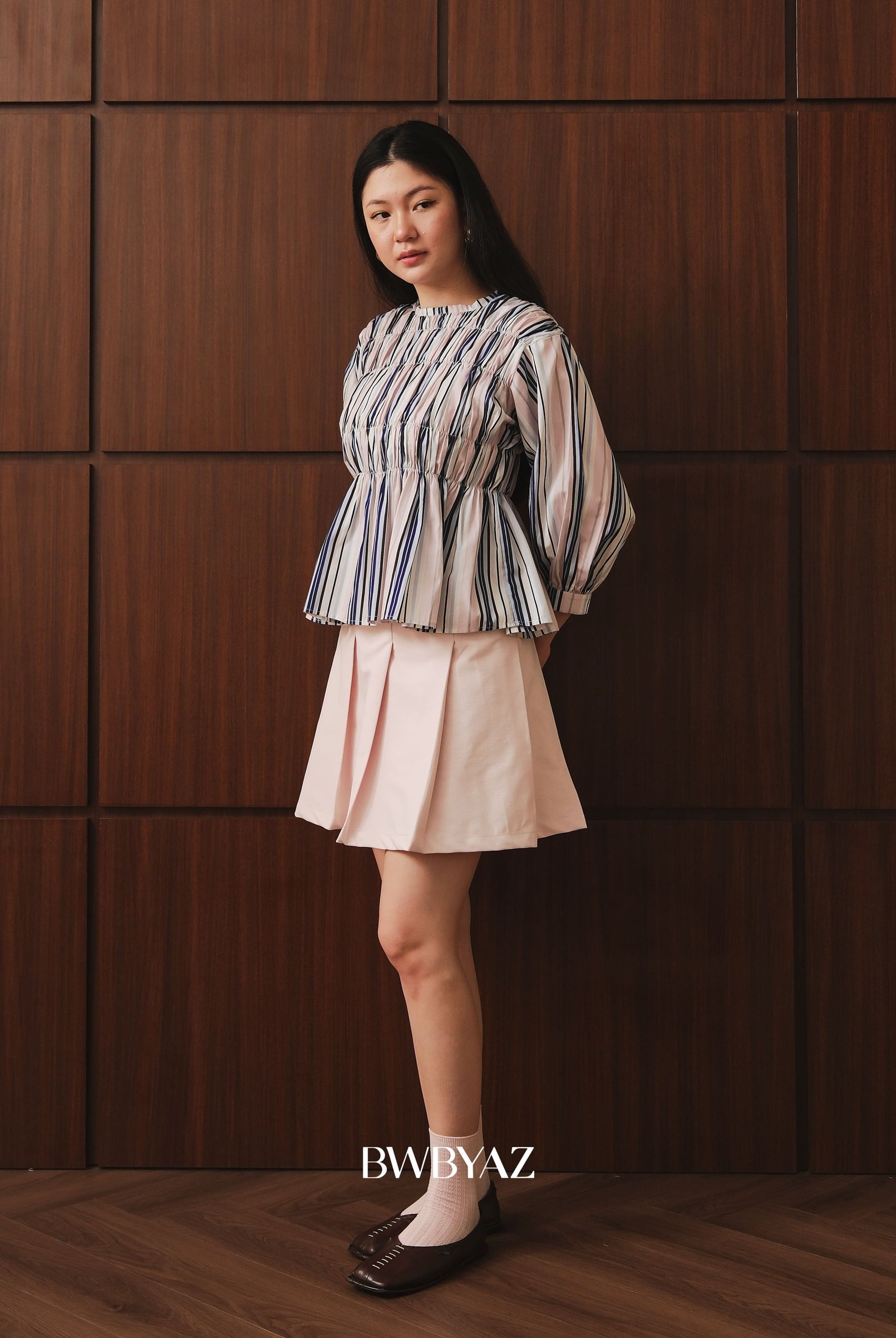 A woman stands against a dark brown wood-paneled wall wearing a striped long-sleeve peplum top by BWBYAZ and a light pink pleated mini skirt. She is wearing white socks and dark brown flat shoes.
