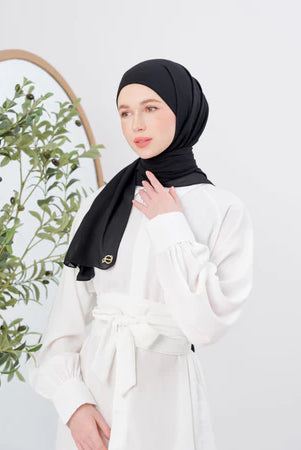 Woman wearing a black pashmina hijab and white outfit with a plant in the background