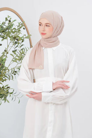 Woman wearing a beige pashmina hijab and white outfit with a plant in the background