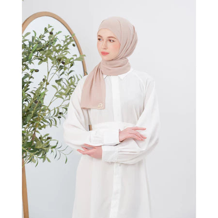 Woman wearing a beige pashmina hijab and white outfit with a plant in the background