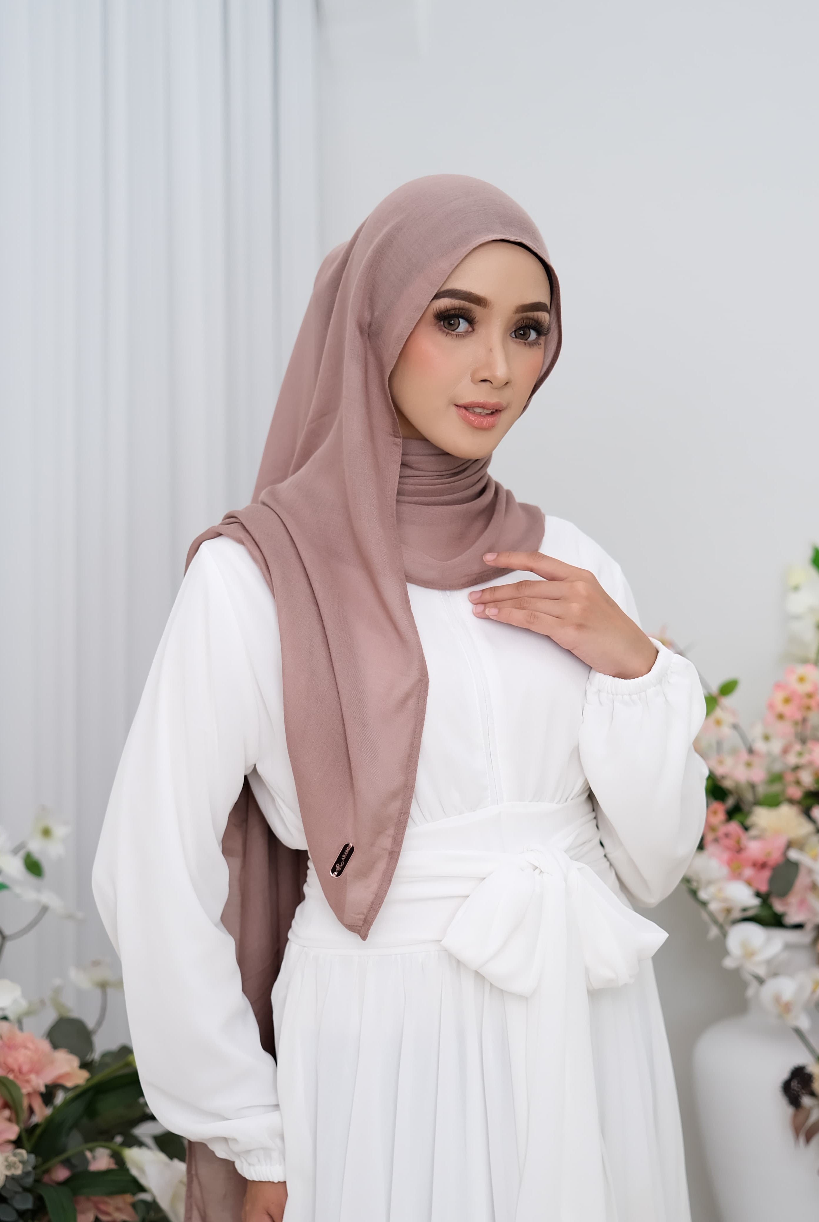 Woman wearing a white dress and brown hijab pashmina viscose with floral decorations in the background