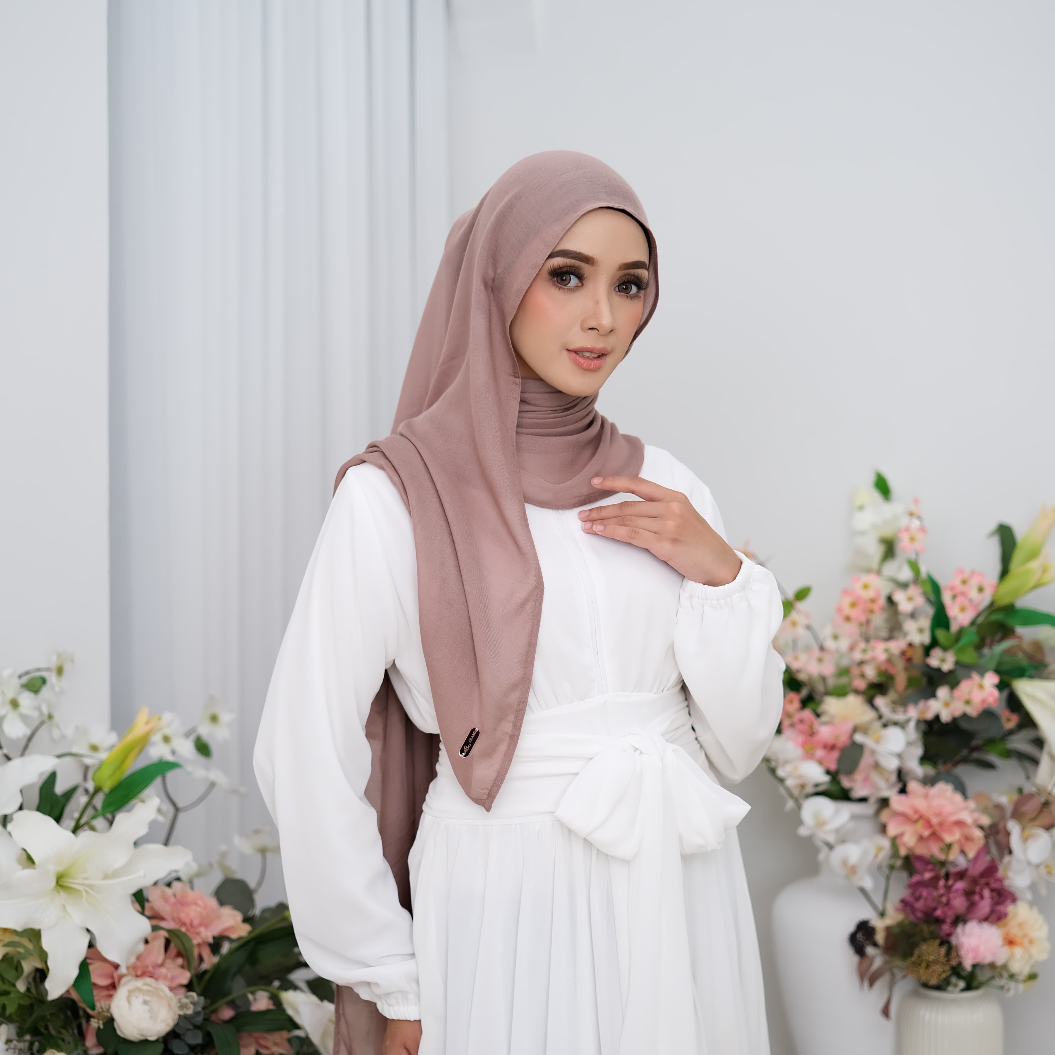 Woman wearing a white dress and brown hijab pashmina viscose with floral decorations in the background