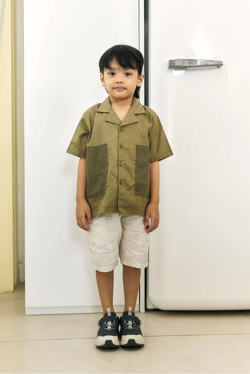 A young boy stands in front of a white refrigerator wearing a short-sleeve olive green button-up shirt by Studio Tui with textured front panels, off-white shorts, and dark sneakers.